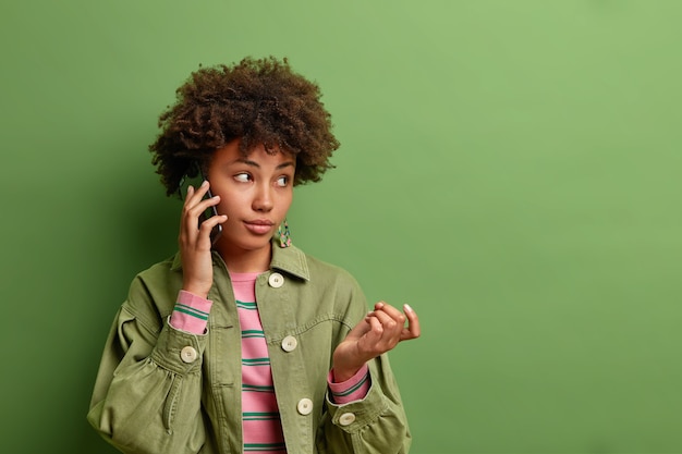 Задумчивая молодая афроамериканка поднимает руку во время телефонного разговора, рассматривает информацию, услышанную от собеседника, носит стильную одежду, изолированную над ярко-зеленой стеной