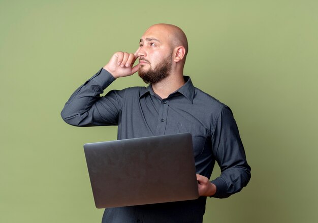 Thoughtful young bald call center man holding laptop looking at side with finger on temple isolated on olive green wall