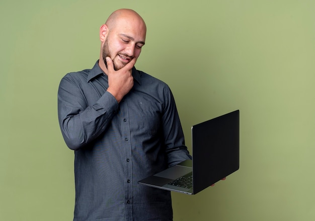 Thoughtful young bald call center man holding and looking at laptop putting hand on chin isolated on olive green wall