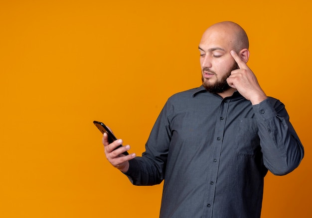 Thoughtful young bald call center man holding and looking at mobile phone putting finger on temple isolated on orange wall