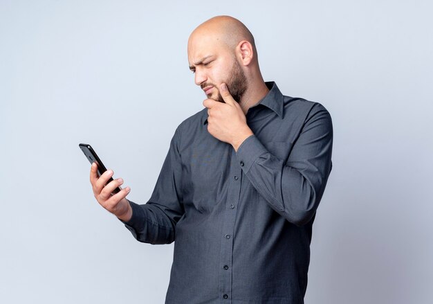 Thoughtful young bald call center man holding and looking at mobile phone with hand on chin isolated on white wall