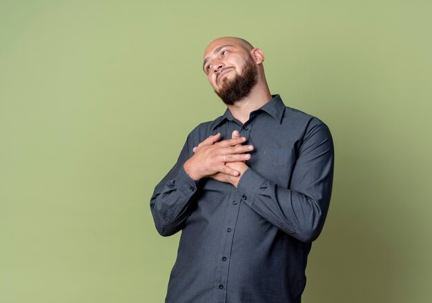 Thoughtful young bald call center man looking straight with hands on chest isolated on olive green wall