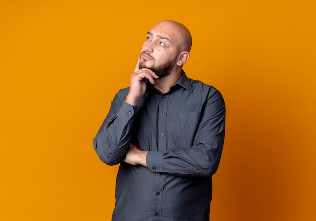 Thoughtful young bald call center man putting hand on chin and looking at side isolated on orange wall