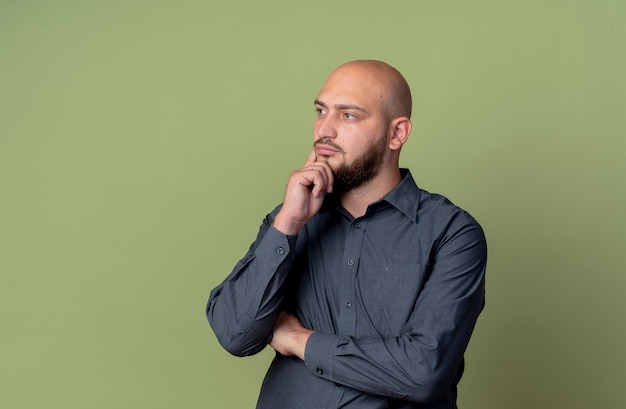 Thoughtful young bald call center man standing with closed posture looking straight with hand on chin isolated on olive green wall