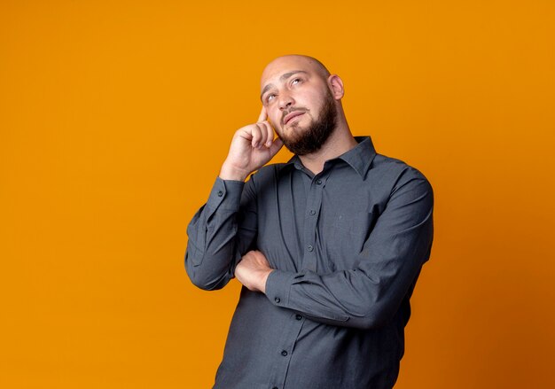 Thoughtful young bald call center man standing with closed posture putting finger on temple looking up isolated on orange wall