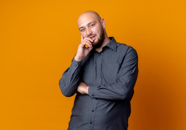 Thoughtful young bald call center man standing with closed posture and putting hand on chin isolated on orange wall