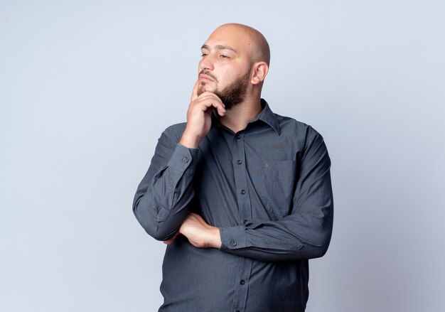 Thoughtful young bald call center man standing with closed posture putting hand on chin looking at side isolated on white wall