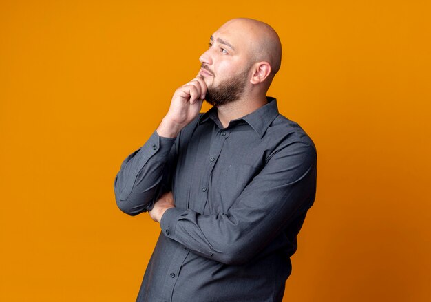 Thoughtful young bald call center man standing with closed posture putting hand on chin looking up isolated on orange wall