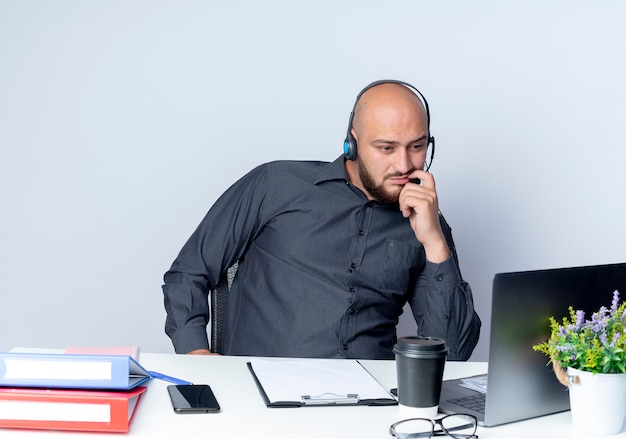 Thoughtful young bald call center man wearing headset sitting at desk with work tools looking at laptop isolated on white wall