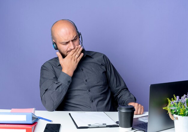 Thoughtful young bald call center man wearing headset sitting at desk with work tools looking at laptop with hand on mouth isolated on purple wall