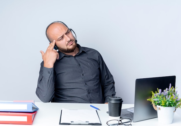 Thoughtful young bald call center man wearing headset sitting at desk with work tools looking at side and putting fingers on temple isolated on white wall