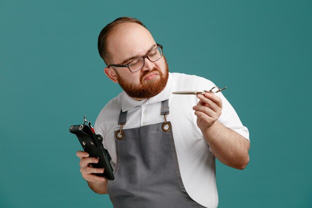 Thoughtful young barber wearing uniform and glasses holding barber bag with barbering tools and scissors looking at scissors isolated on blue background