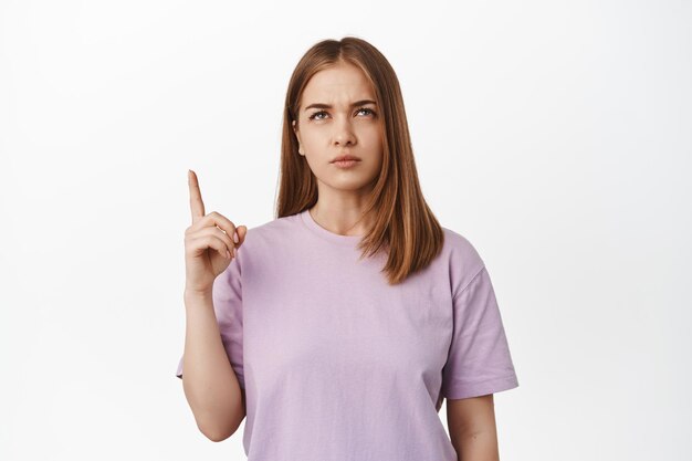 Thoughtful young blond woman, girl looks and points up with disbelief, squinting pensive, thinking, making decision, feeling unsure about logo text, standing against white background