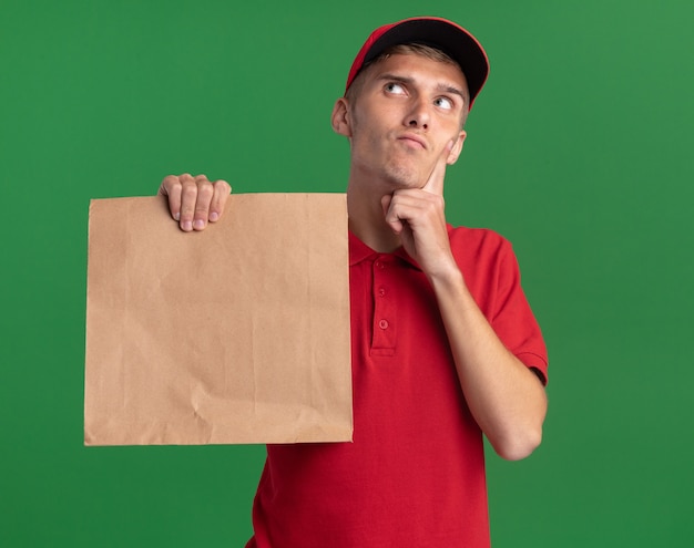 Thoughtful young blonde delivery boy puts finger on chin and holds paper package looking up isolated on green wall with copy space