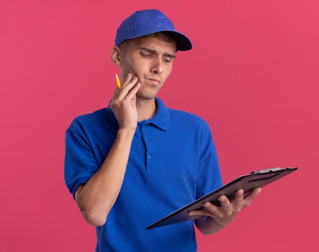 Thoughtful young blonde delivery boy puts hand on face holding and looking at clipboard isolated on pink wall with copy space