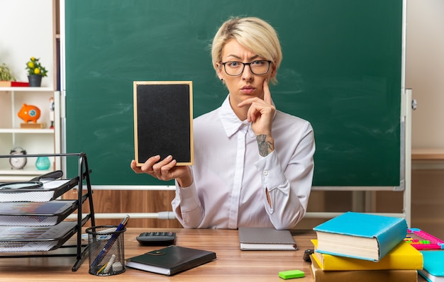 thoughtful young blonde female teacher wearing glasses sitting at desk with school supplies in classroom showing mini blackboard keeping hand on chin looking at front