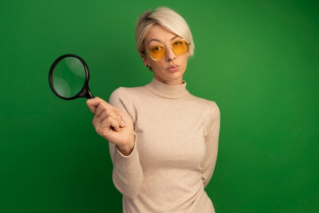 Thoughtful young blonde girl wearing sunglasses holding magnifying glass keeping hand behind back 