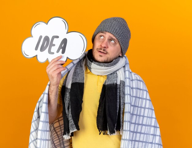 Thoughtful young blonde ill slavic man wearing winter hat and scarf wrapped in plaid holds idea bubble looking up isolated on orange wall with copy space