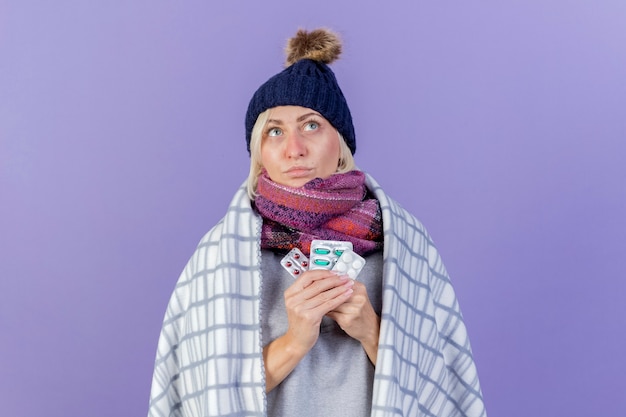 Thoughtful young blonde ill slavic woman wearing winter hat and scarf wrapped in plaid holds packs of medical pills looking up isolated on purple wall with copy space