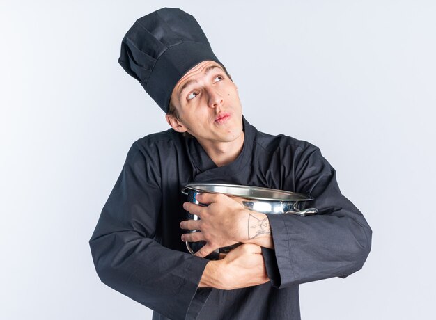 Thoughtful young blonde male cook in chef uniform and cap holding and hugging pot looking up isolated on white wall