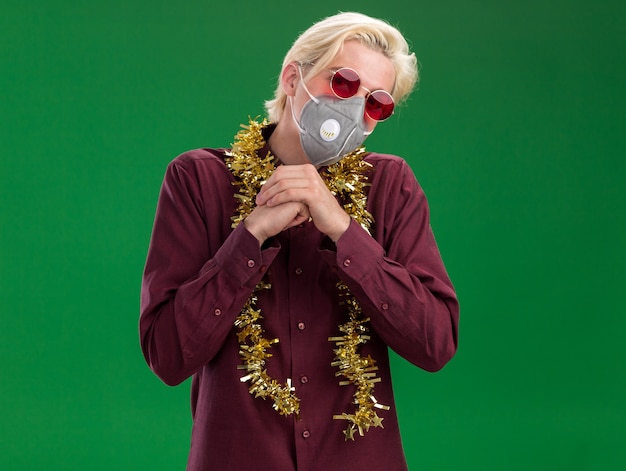 Free photo thoughtful young blonde man wearing glasses and protective mask with tinsel garland around neck  keeping hands together isolated on green wall with copy space