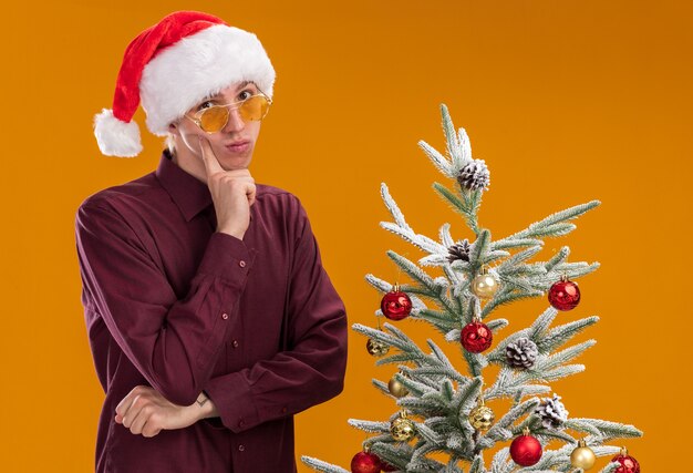 Thoughtful young blonde man wearing santa hat and glasses standing near decorated christmas tree on orange background