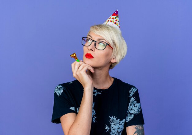 Задумчивая молодая блондинка тусовщица в очках и кепке на день рождения, держащая воздуходувку, смотрящую в сторону, изолированную на фиолетовом фоне с копией пространства