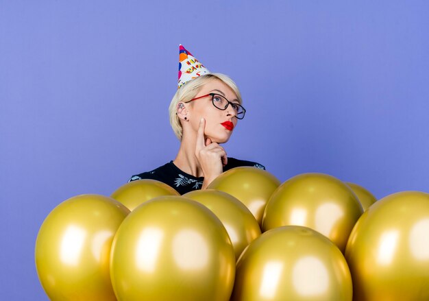 Thoughtful young blonde party girl wearing glasses and birthday cap standing behind balloons looking up touching face isolated on purple background