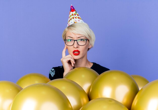 Thoughtful young blonde party girl wearing glasses and birthday cap standing behind balloons touching face looking at camera isolated on purple background