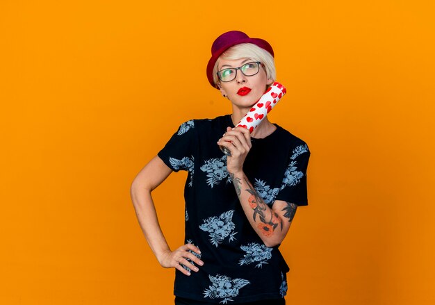 Thoughtful young blonde party girl wearing party hat and glasses keeping hand on waist looking at side touching face with confetti cannon isolated on orange background with copy space