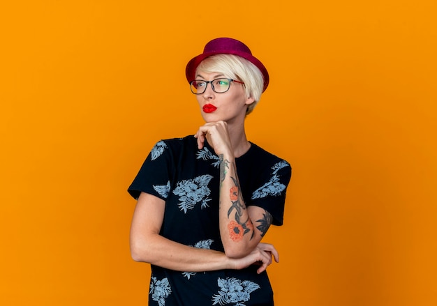 Thoughtful young blonde party girl wearing party hat and glasses putting hand under chin looking at side isolated on orange background with copy space