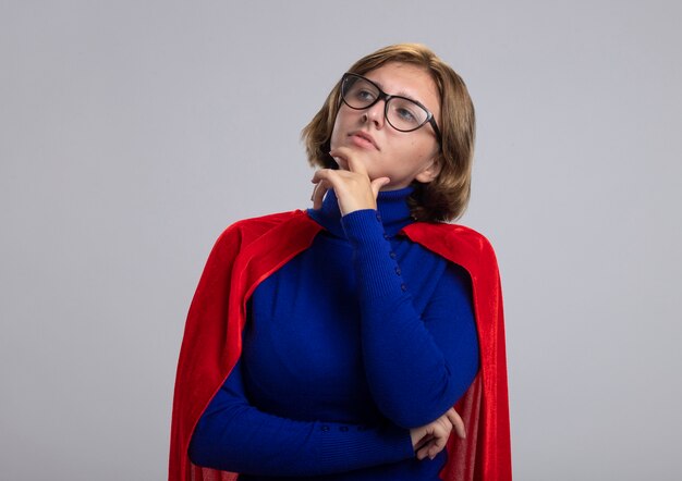 Thoughtful young blonde superhero girl in red cape wearing glasses touching chin looking at side isolated on white background