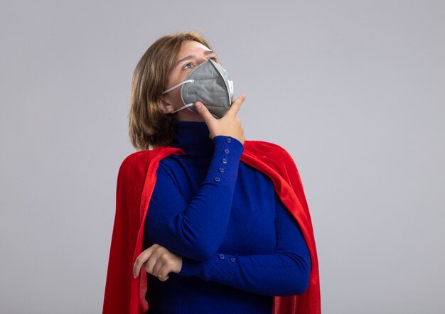 Thoughtful young blonde superhero girl in red cape wearing protective mask looking up touching up isolated on white background with copy space