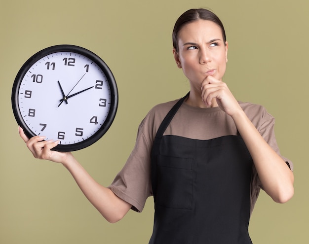 Thoughtful young brunette barber girl in uniform puts hand on chin and holds clock looking up isolated on olive green wall with copy space