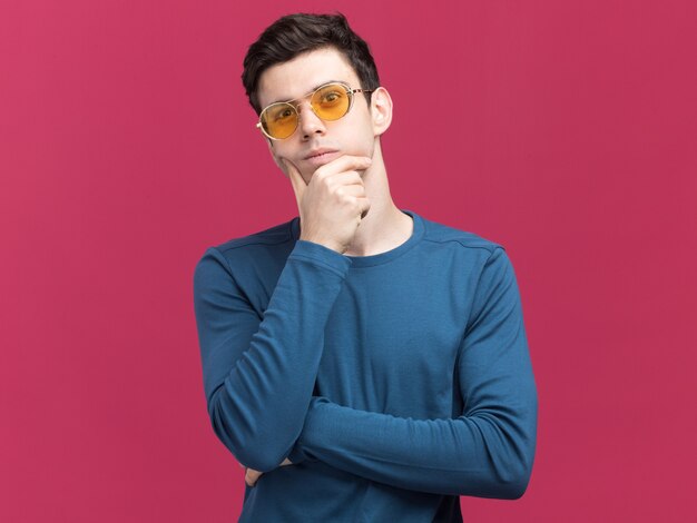 thoughtful young brunette caucasian boy in sun glasses puts hand on chin on pink