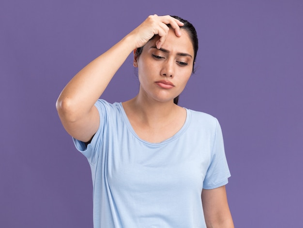 Thoughtful young brunette caucasian girl puts hand on head and looks down isolated on purple wall with copy space