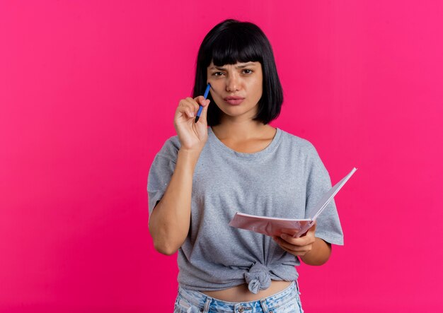 Thoughtful young brunette caucasian girl puts pen on face and holds notebook looking at camera isolated on pink background with copy space