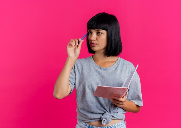 La giovane donna caucasica castana premurosa mette la penna sul tempiale e tiene il taccuino che esamina il lato isolato su fondo rosa con lo spazio della copia