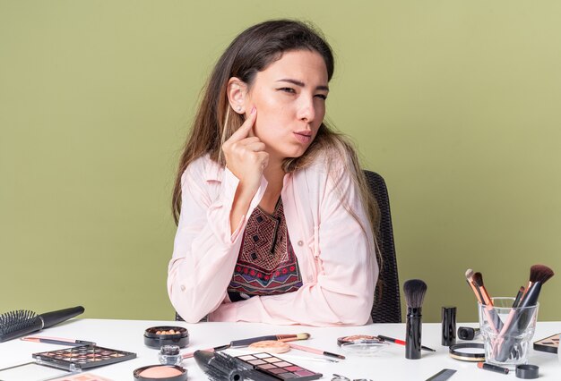 Thoughtful young brunette girl sitting at table with makeup tools looking at side 