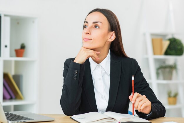 Thoughtful young businesswoman holding pencil on diary over the desk