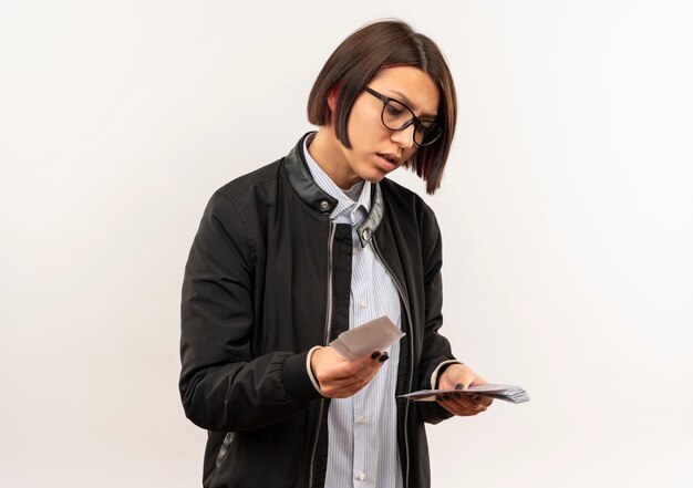 Thoughtful young call center girl wearing glasses holding and looking at money isolated on white wall