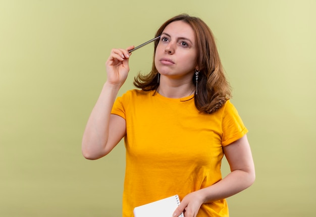 Thoughtful young casual woman holding pen and note pad on isolated green wall with copy space