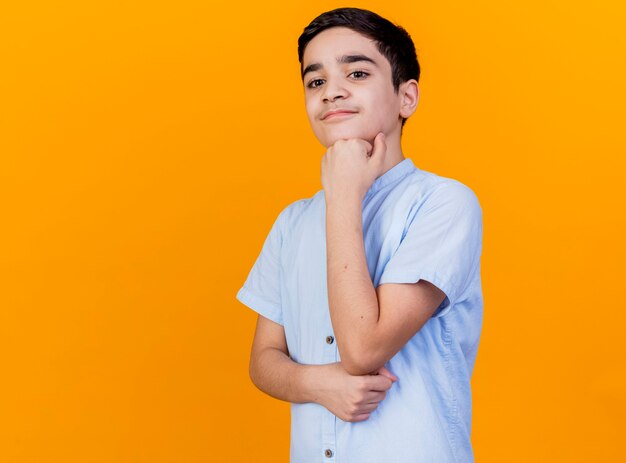 Thoughtful young caucasian boy putting hand under chin looking down isolated on orange background with copy space