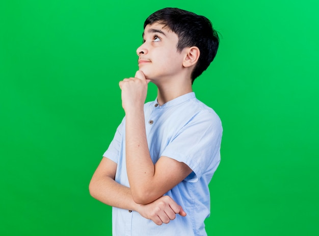 Thoughtful young caucasian boy standing in profile view touching chin looking up isolated on green wall with copy space