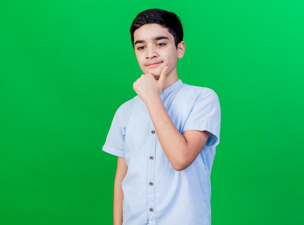 Thoughtful young caucasian boy touching chin looking down isolated on green wall with copy space