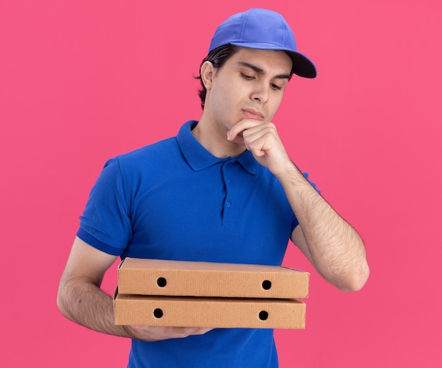 Вдумчивый молодой кавказский доставщик в синей форме и кепке держит и смотрит на пакеты с пиццей, держа руку на подбородке, изолированном на розовой стене