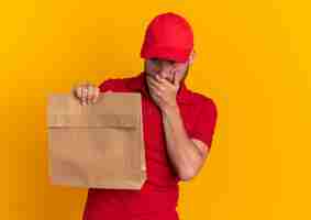 Foto gratuita giovane fattorino caucasico premuroso in uniforme rossa e cappuccio che tiene il pacchetto di carta tenendo la mano sulla bocca guardando in basso