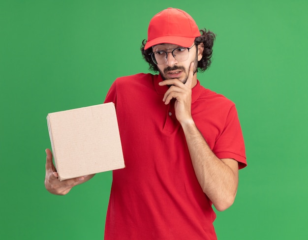 赤い制服を着た思いやりのある若い白人配達人と緑の壁で隔離されたあごに手を保持しているカードボックスを保持し、見ている眼鏡をかけているキャップ