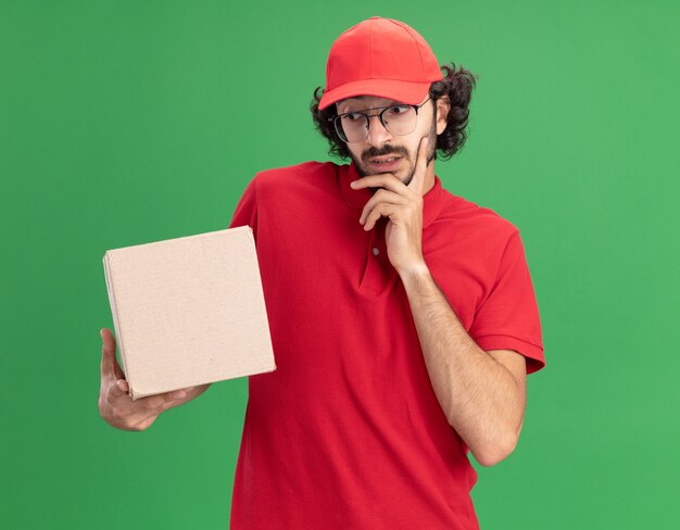 사려깊은 젊은 백인 배달원은 빨간 제복을 입고 안경을 쓰고 녹색 벽에 격리된 턱에 손을 대고 있는 카드박스를 보고 있다
