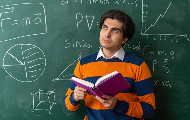 Thoughtful young caucasian geometry teacher standing in front of chalkboard in classroom holding book looking up